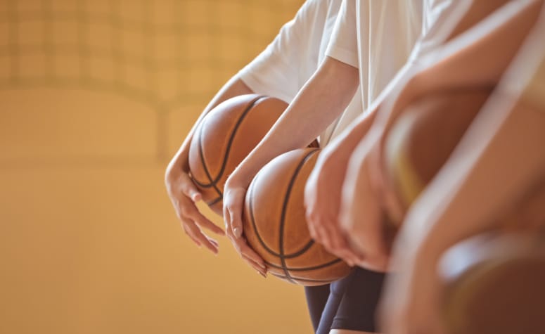 Group of teenager wearing white t-shirts and black shorts standing in row in gym and holding basketball balls. Close up of hands, unrecognizable people.